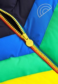 Viacfarebná džínsová bunda so sekciami modrej, zelenej, žltej a čiernej. Žltý zips s dúhovými detailmi a logo na látke.