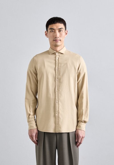 Camicetta lunga beige chiaro con una patta di bottoni e una texture morbida. Abbinata a pantaloni grigi svasati e sartoriali. Design del colletto semplice.