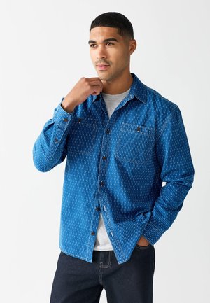 Homme portant une chemise bleue à motifs boutonnée sur un t-shirt gris, une main dans la poche et l'autre ajustant le col, debout sur fond blanc.