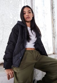 Vrouw met lang donker haar, gekleed in een zwarte doorgestikte jas, een witte cropped top en olijfgroene broek, zittend tegen een gestructureerde muur.