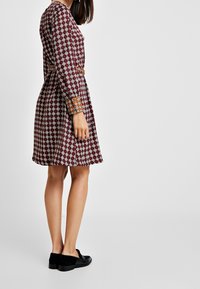 Robe bordeaux avec un motif géométrique blanc, manches longues, taille cintrée et accents orange contrastants sur les manches. Des chaussures noires complètent la tenue.