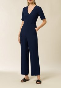 Jumpsuit blu navy con maniche corte, scollo a V e vita aderente. Progettato con gambe larghe e dritte e tasche laterali. Sandali piatti.