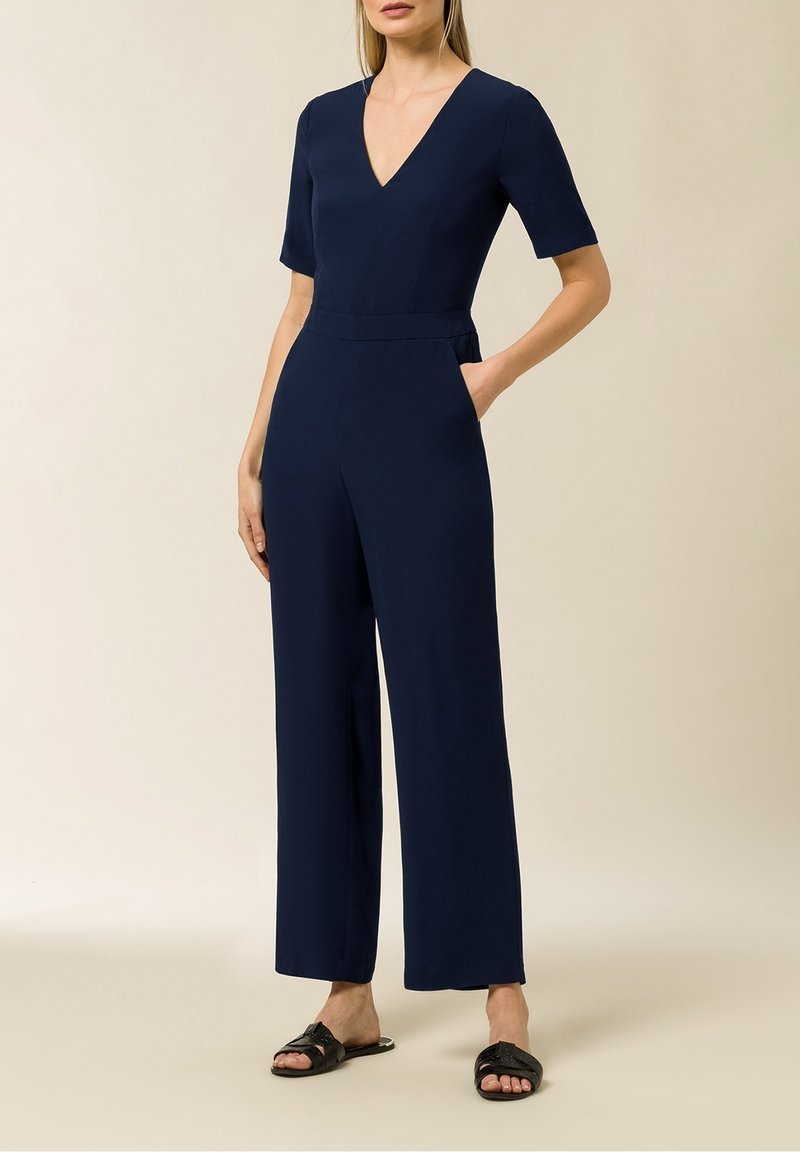 Jumpsuit blu navy con maniche corte, scollo a V e vita aderente. Progettato con gambe larghe e dritte e tasche laterali. Sandali piatti.