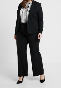 Veste de costume noire sur une chemise blanche à rayures verticales noires, assortie à un pantalon large noir et des escarpins noirs.