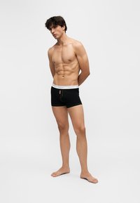 Jeune homme torse nu, debout pieds nus, portant un boxer noir avec une ceinture blanche sur un fond clair uni.