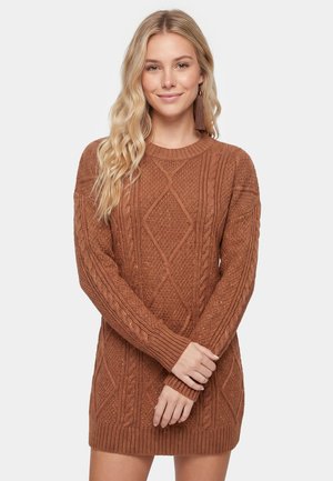 Young woman with long blonde hair wearing a brown cable knit sweater dress and long tassel earrings, posing with hands crossed.