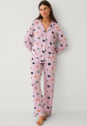 Woman wearing light purple button-up pajama set with colorful sushi print, standing barefoot against a plain light gray background.