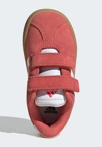 Červené semišové tenisky s dvoma popruhmi na suchý zips, bielymi akcentami a polstrovanou vložkou. Obsahujú zaoblenú špičku a béžovú gumovú podrážku.