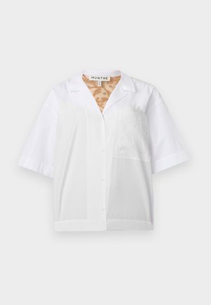 Kvinne står med hånden i lommen, iført løs hvit kortermet skjorte, brede mørke jeans og brune sko på ensfarget bakgrunn.