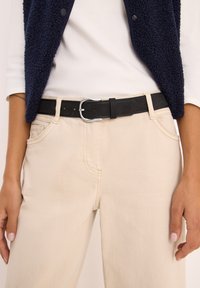 Zwarte riem met een zilveren gesp, gedragen over lichtbeige broek. Het materiaal lijkt op leer, met een gladde textuur en meerdere verstelgaatjes.