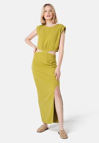 Vestido ajustado hasta la rodilla en verde olivo, con acentos cortados, mangas cortas y abertura lateral; combinado con sandalias planas beige.
