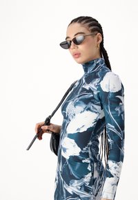 Μακρυμάνικο, εφαρμοστό φόρεμα με λουλουδάτο μπλε και άσπρο σχέδιο, φτιαγμένο από λεία ύφασμα. Τα αξεσουάρ περιλαμβάνουν μαύρα γυαλιά ηλίου σε σχήμα γάτας και μια μικρή τσάντα.