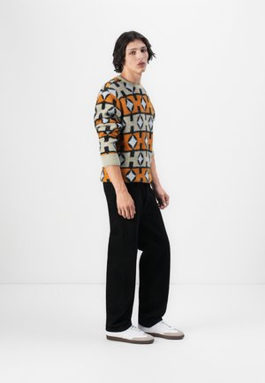 Knit sweater in beige with orange, black, and gray geometric patterns; paired with black pants and white shoes featuring gray accents.