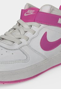 Λευκό αθλητικό παπούτσι Nike με ροζ λουράκι, ροζ λογότυπο Nike Swoosh, λευκά κορδόνια και ροζ εξωτερική σόλα, ορατά σε ανοιχτό φόντο.