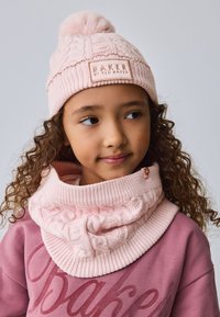 Roza pletena kapa s cofom in logotipno značko, v kombinaciji z ujemajočim se puloverjem z visokim ovratnikom in teksturiranim vzorcem.