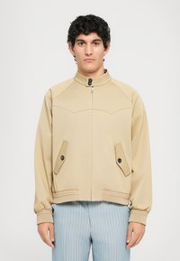 Moški v bež jopiču na zadrgo z gumbi in modrih prugastih hlačah, stoji pred enostavno svetlo ozadjem.