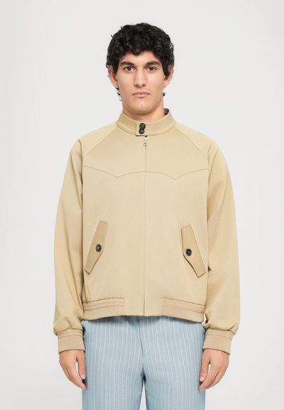 Mees, kes kannab beeži tõmbluku ja nööpidega taskutega jope ning siniseid triibulisi pükse, seisab ühetaolise heleda tausta ees.