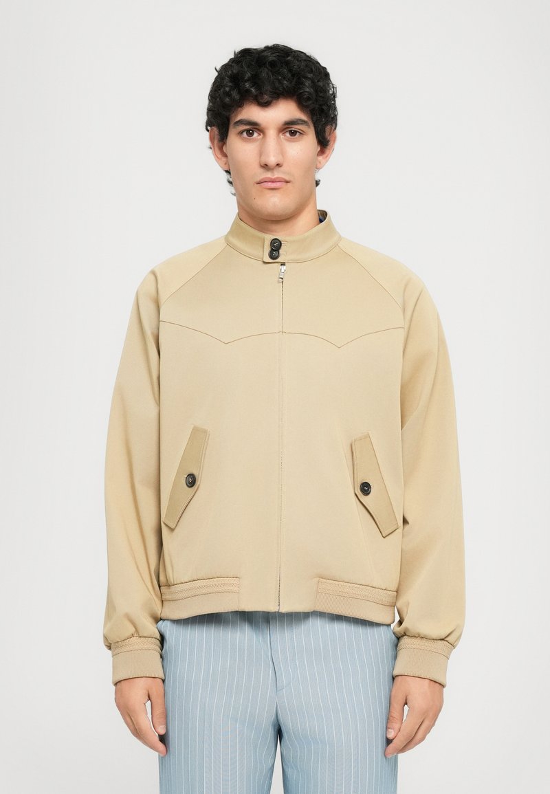 Man wearing beige zip-up jacket with buttoned collar and flap pockets, paired with light blue pinstripe pants, standing against white background.