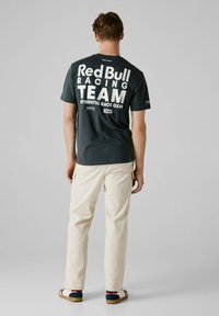 Mørkegrå t-shirt med stort hvidt tekst på ryggen, der står "Red Bull Racing TEAM," parret med lyse beige bukser og casual sko.