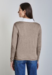 Bruine gebreide trui met een ronde halslijn, lange mouwen en een lichtblauw overhemd eronder. Gecombineerd met blauwe spijkerbroek.