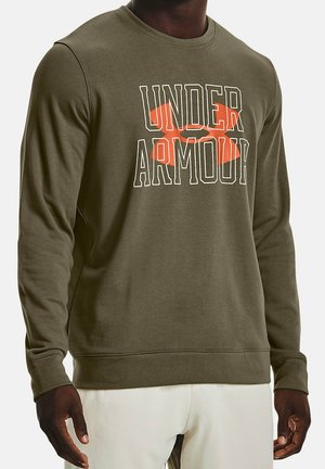 Mand iført olivengrøn langærmet sweatshirt med stort orange og hvidt "Under Armour" logo foran, kombineret med hvide bukser.
