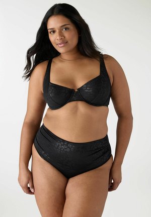 Donna con lunghi capelli scuri che indossa un reggiseno nero con ferretto e motivo stampato e slip bikini a vita alta contro uno sfondo bianco uniforme.