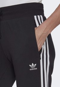 Mustat Adidas-housut valkoisilla raidoilla, joissa on kiristettävä vyötärö ja sivu-zip-tasku. Ikoninen logo alhaalla sääressä. Sujuva pinta.