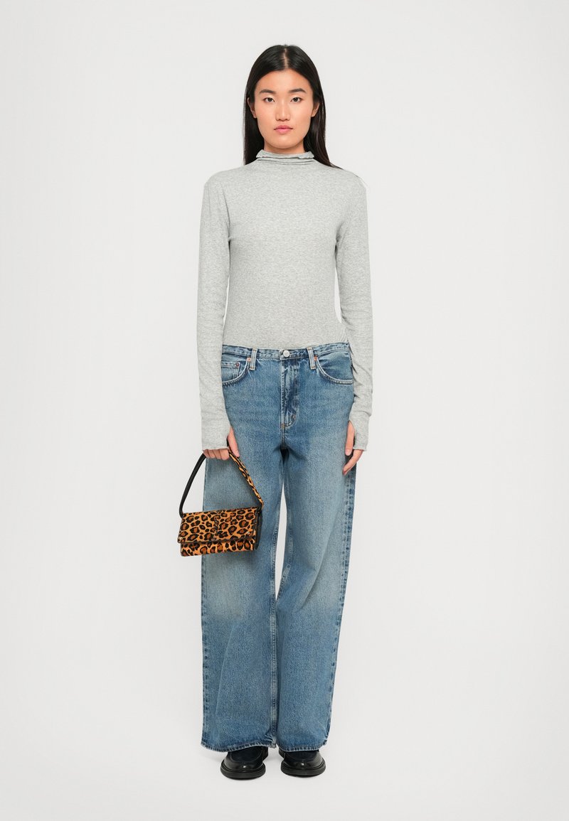 Femme portant un haut gris à manches longues, un jean bleu à jambes larges, des chaussures noires et tenant un petit sac à main en imprimé léopard devant un fond uni.