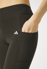 Leggings deportivos negros con una textura lisa, que cuentan con un bolsillo lateral y un detalle de logo de tres rayas blancas cerca de la cinturilla.