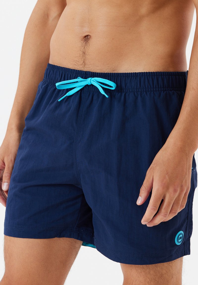 Bañadores de color navy con cintura elástica y cordón turquesa. Cuentan con un bolsillo lateral y un logo circular en el dobladillo. Textura de tela suave.