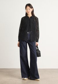 Black lace shirt with button closure and two chest pockets, paired with wide-leg dark denim jeans and silver-tone shoes. Small black handbag.