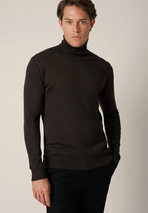 Mężczyzna w ciemnobrązowym prążkowanym golfie i czarnych spodniach, stojący z jedną ręką w kieszeni na neutralnym tle.