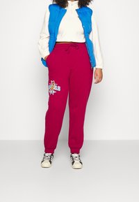 En person som bär en blå pufferväst, en vit kortärmad topp, magenta svettbynor med tryck av tecknade hjortar och svart-vita sneakers, står med händerna i fickorna.