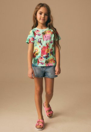 Junges Mädchen mit langen Haaren, das ein buntes T-Shirt mit tropischem Muster, Jeansshorts und rosa Sandalen trägt, vor einem schlichten beigen Hintergrund steht.