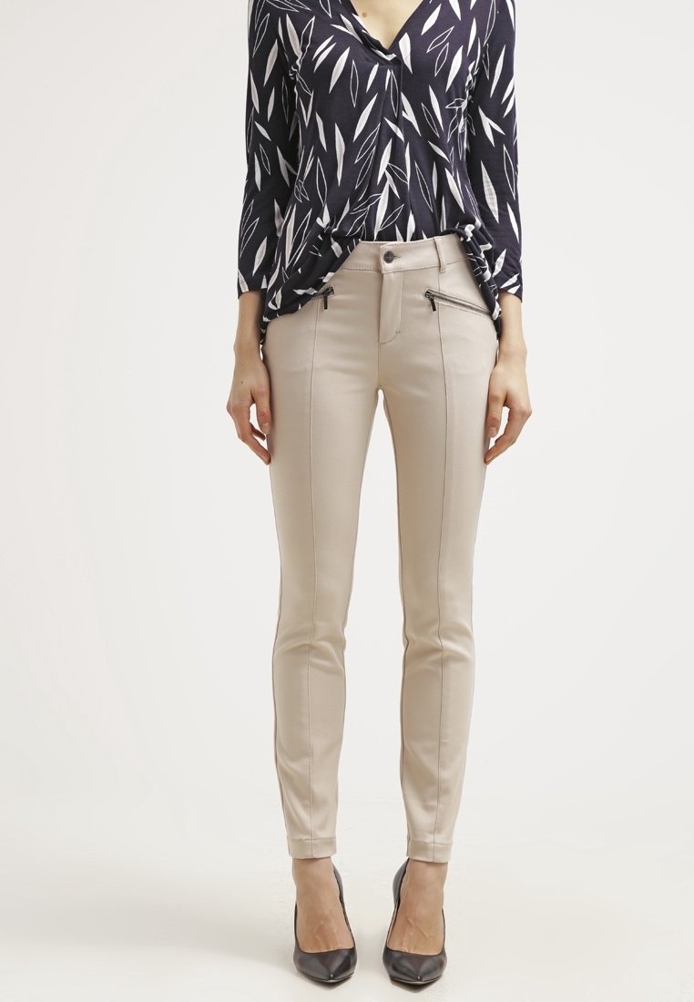 Femme portant un pantalon beige ajusté avec une couture avant et des poches zippées, associé à un chemisier à motifs noir et blanc et des talons noirs.