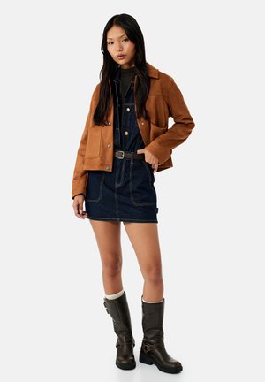 Młoda kobieta ubrana w brązową zamszową kurtkę, ciemną sukienkę dżinsową z paskiem oraz czarne kozaki za kolano z białymi skarpetkami, stojąca na jednolitym tle.