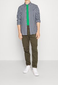 Camisa de cuadros en azul marino y blanco, llevada sobre una camiseta verde, combinada con pantalones verde oliva y zapatillas blancas.