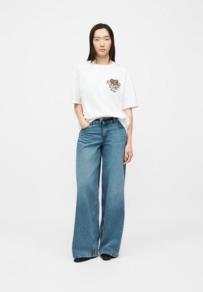 Femme aux cheveux noirs et raides, debout, portant un t-shirt blanc à motif graphique et un jean bleu à jambes larges avec des chaussures noires, main dans la poche.