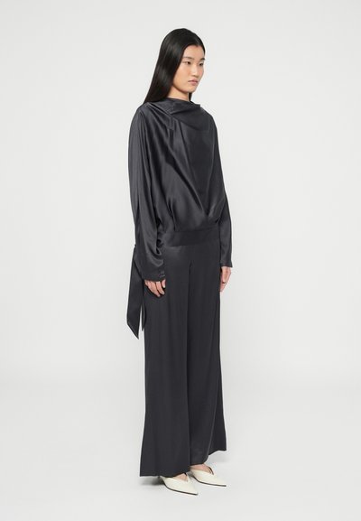 Femeie cu păr lung și negru, purtând o bluză largă de mătase neagră și pantaloni evazați, cu pantofi albi cu vârf ascuțit, stând pe un fundal alb.