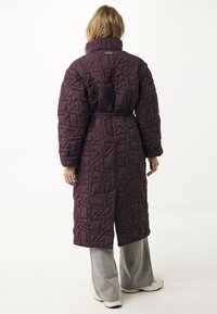 Donna che indossa un lungo cappotto trapuntato di un viola scuro con colletto alto, vita cintrata, abbinato a pantaloni grigi chiari a gamba larga e sneaker bianche.