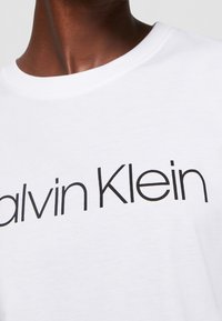 White cotton t-shirt featuring the "Calvin Klein" logo in bold black lettering, round neckline, and short sleeves. Smooth texture.