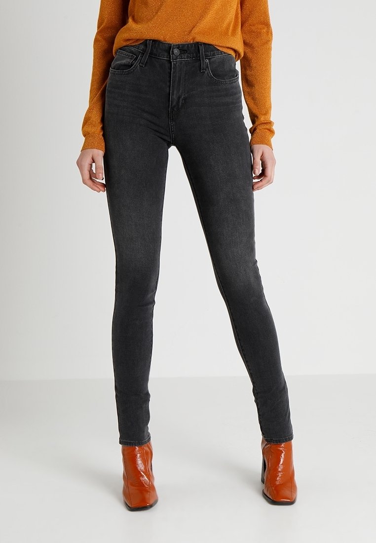 Jeans negros ajustados de denim con cintura alta, que presentan un diseño clásico de cinco bolsillos y detalles de costura mínimos, combinados con una blusa naranja y botines.