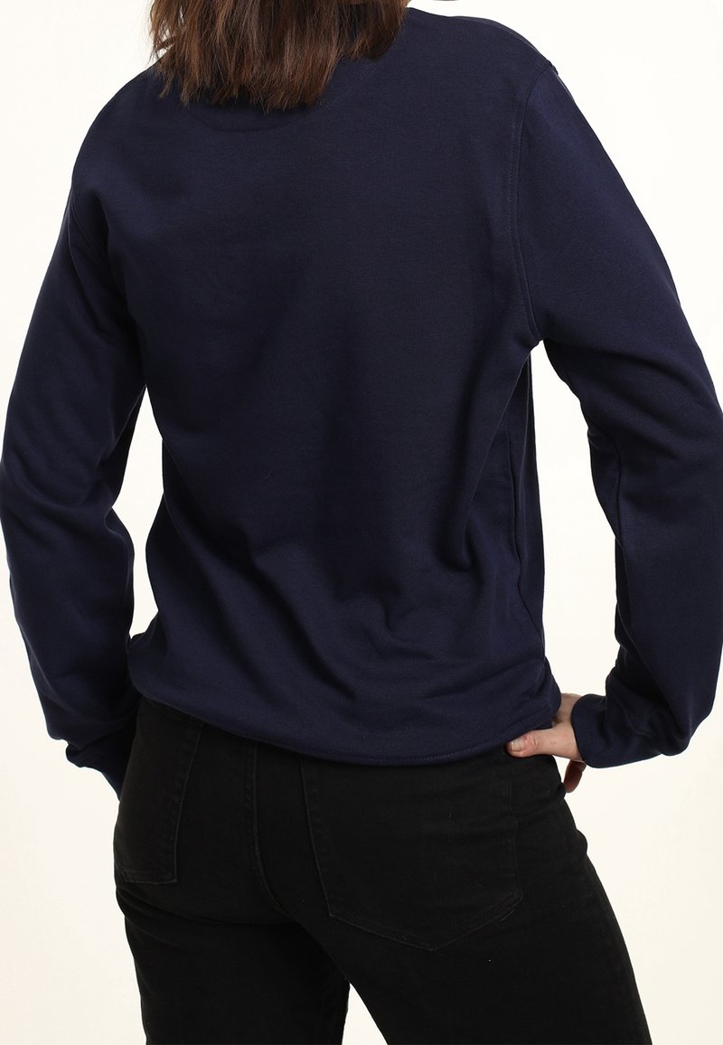 Person wearing a navy blue long-sleeve shirt and black pants, standing with one hand on their hip, facing away from the camera.