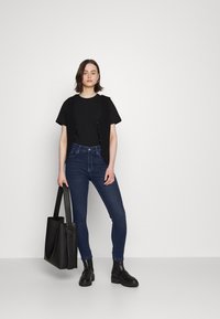 Colete preto de malha sobre uma t-shirt preta, combinado com jeans skinny azul-escuro e botas de plataforma pretas. Acessorizado com uma bolsa tote preta.