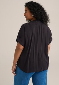 Čierny top s krátkym rukávom a štruktúrovanou tkaninou. Má zaoblený lem a jemný zadný záhyb pre dodatočný detail. Skombinovaný s modrými nohavicami.