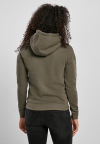 Sweat à capuche vert olive avec un cordon de serrage, des poignets et une ceinture côtelés. Texture lisse avec une coupe décontractée, doté d'une capuche de taille moyenne.