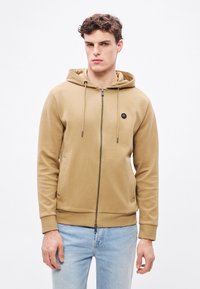 Khaki mikina na zips s kapucňou so šnúrkou, rebrovanými manžetami a kruhovým logom na hrudi. Mäkká tkanina, užší strih, doplnená svetlomodrými džínsami.