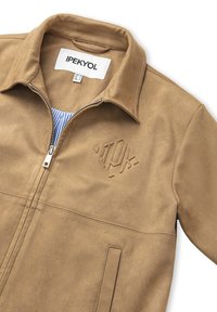 Béžová semišová bunda se zipem, límcem a vyšitým logem. Obsahuje boční kapsy a prošívané detaily, které zdůrazňují její strukturovaný design.