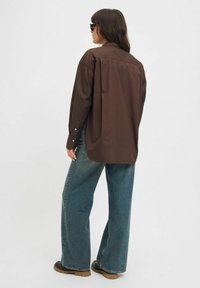 Camicia a collo largo marrone di grandi dimensioni realizzata in tessuto morbido, abbinata a jeans azzurri svasati e scarpe marroni. Aggiunti occhiali da sole.