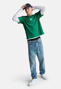 Camiseta verde de manga corta superpuesta sobre una camisa blanca de manga larga, combinada con unos vaqueros anchos de color azul claro y zapatillas blancas. Gorra negra en la cabeza.